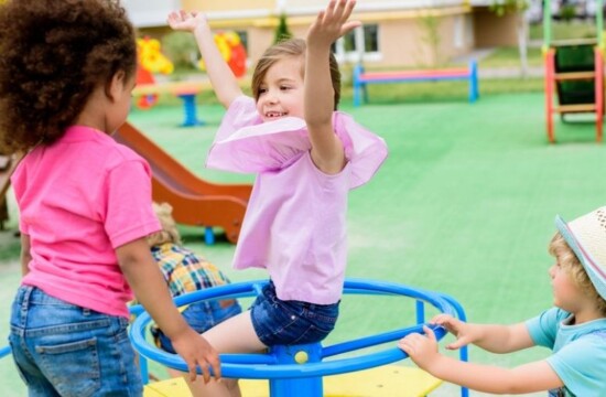 Conheça as vantagens de usar grama sintética para playground com a Stampa Gramas Crianças felizes brincam no gira-gira, que tem o chão seguro de grama sintética para playground.