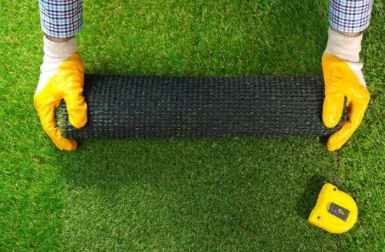 Conheça mitos e verdades sobre o uso da grama sintética! Mãos com luvas amarelas desenrolando um tapete verde sobre o solo, exemplificando o uso da grama sintética.